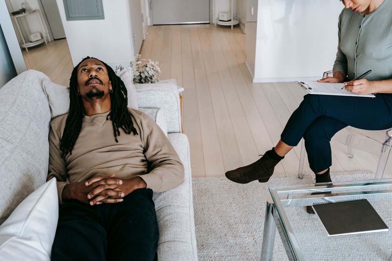 From above of African American male looking up while lying on couch near crop female therapist writing notes on clipboard during session
