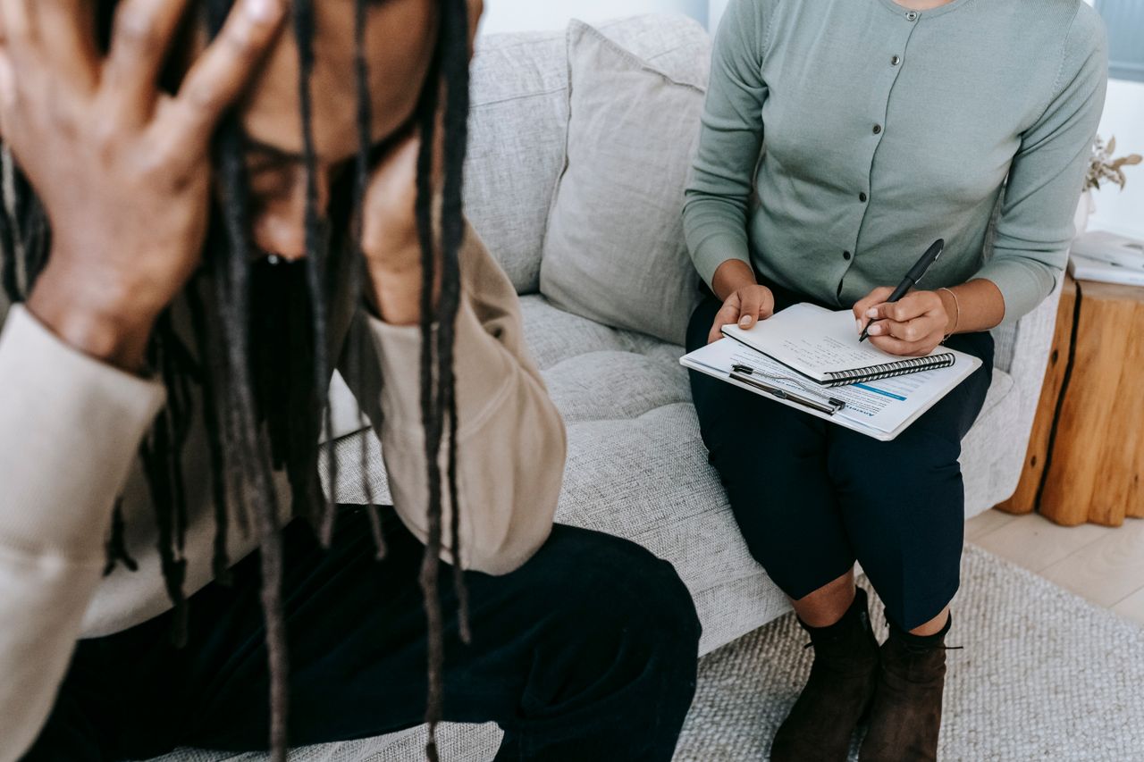 Crop concerned African American male sitting on sofa with female psychologist and holding head in hands in dismay during psychotherapy session