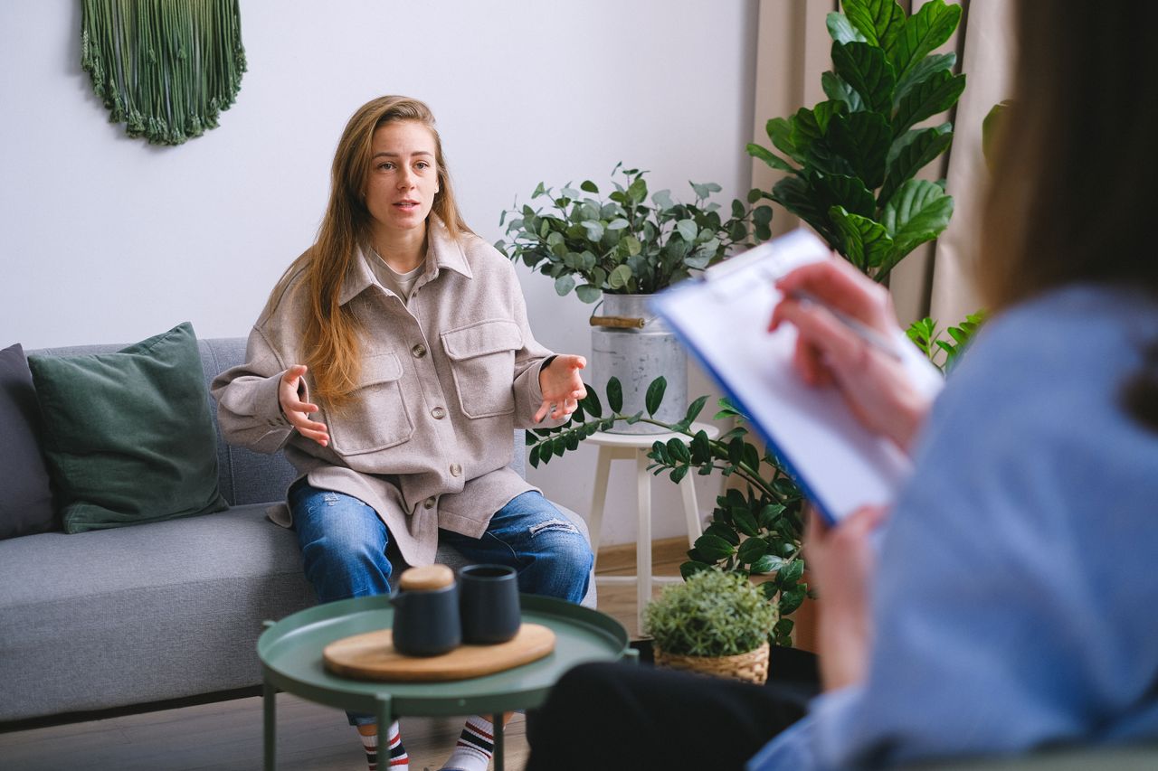 Concerned female client siting on couch and speaking about mental problems during psychotherapy appointment with blurred psychologist in light office